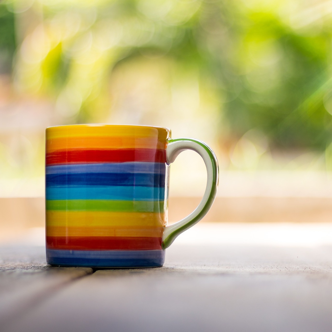 Bunte regenbogenfarbene Tasse mit grünem Henkel auf Holzoberfläche.