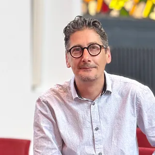 Lächelnder Mann mit Brille und grau meliertem Haar sitzt drinnen.