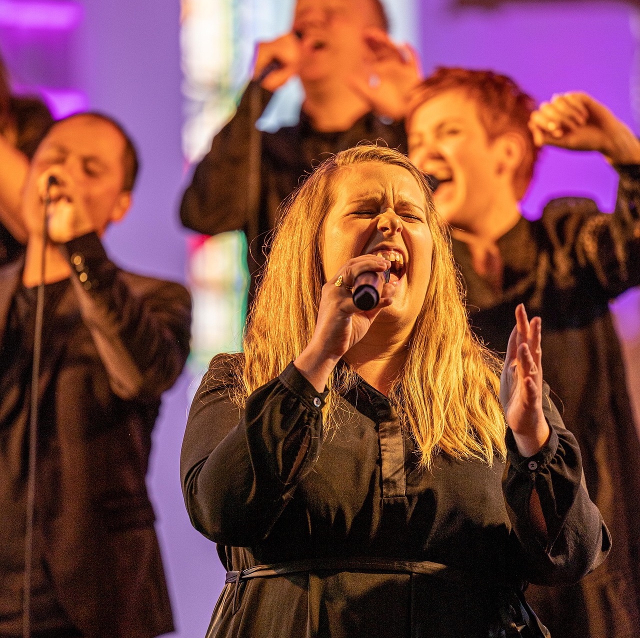 Frau singt leidenschaftlich ins Mikrofon mit Chormitgliedern im Hintergrund.