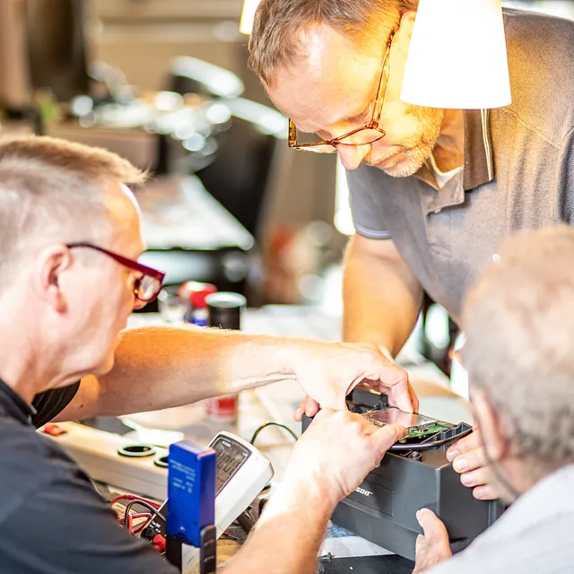 Drei Männer arbeiten gemeinsam an elektronischer Ausrüstung an einem Tisch.