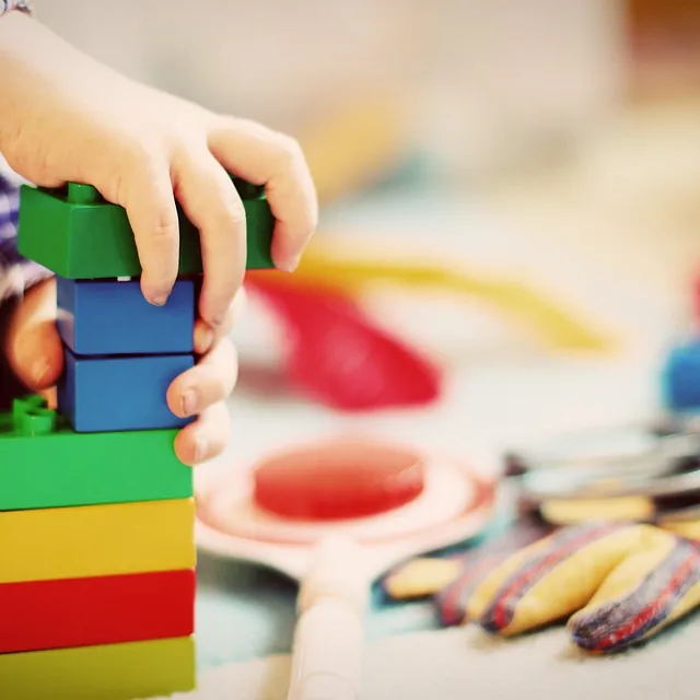 Kinderhände stapeln bunte Bauklötze auf einer Spielfläche.