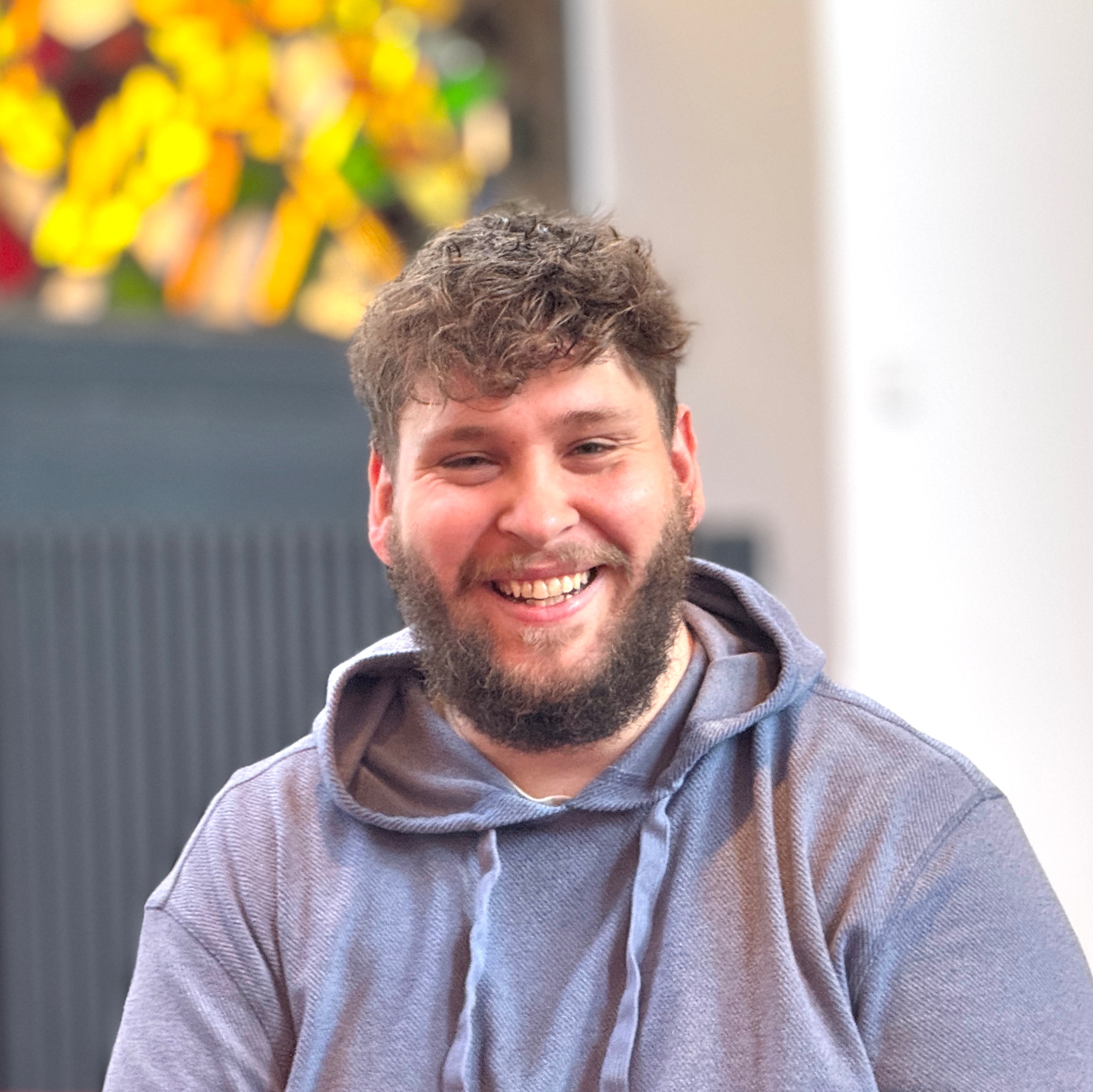 Lächelnder junger Mann mit lockigem Haar und Bart drinnen.