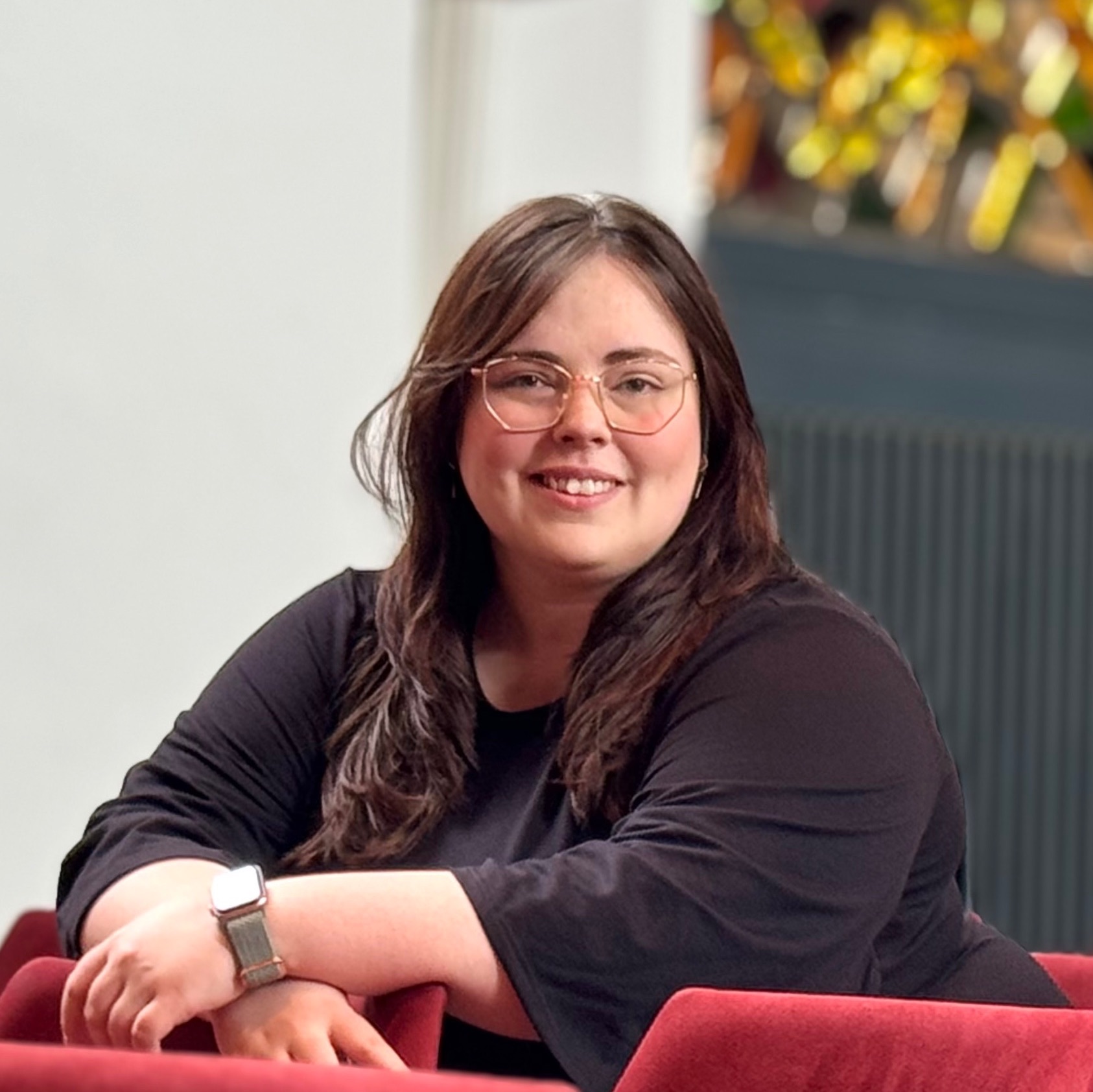 Lächelnde Frau mit Brille und langen dunklen Haaren auf rotem Stuhl.