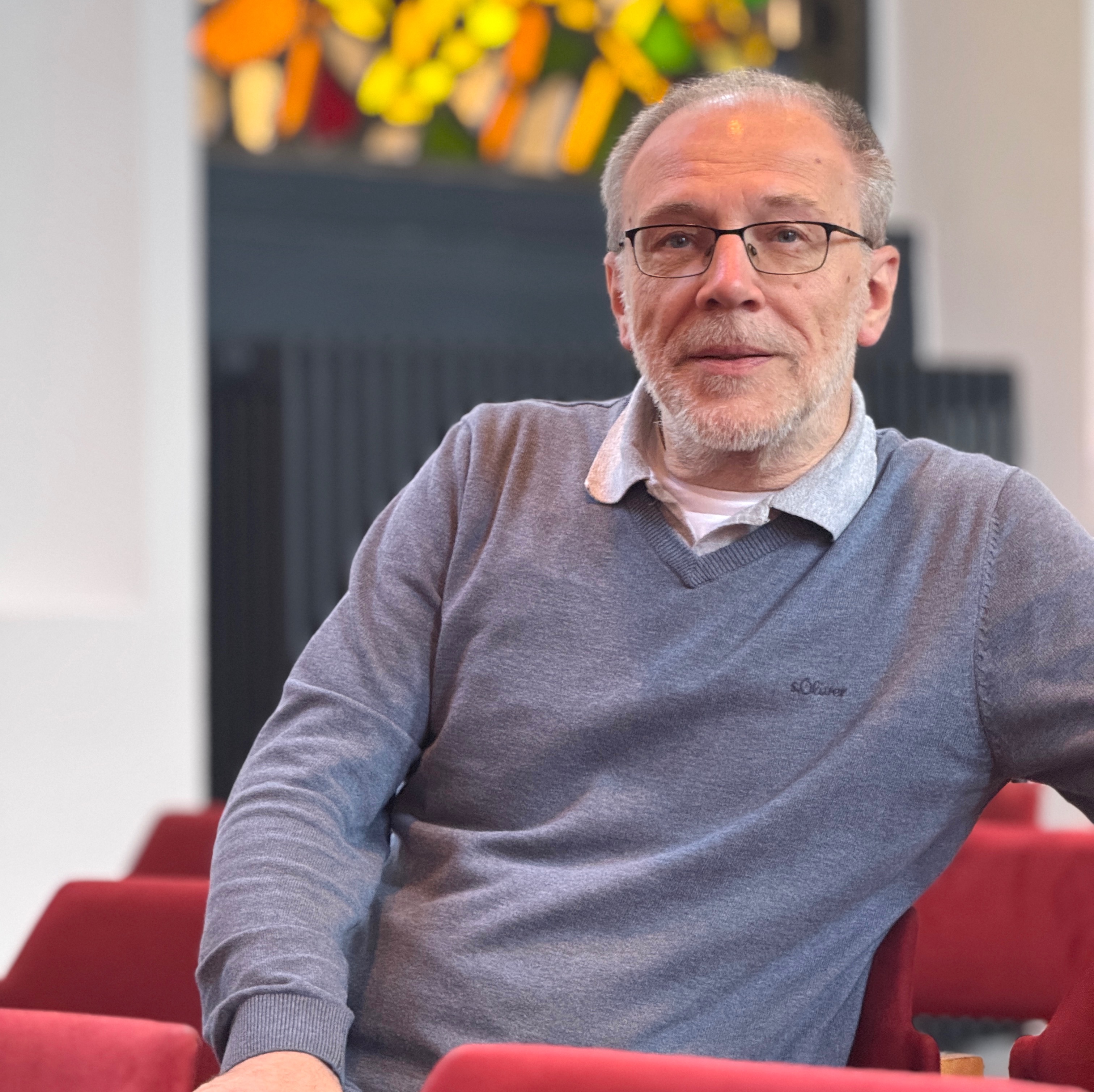  Älterer Mann mit Brille sitzt in roter Kirchenbank und schaut nach vorne.
